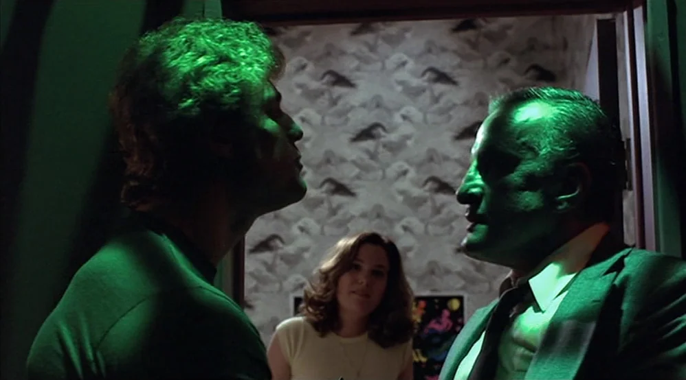 Hardcore (1979) movie still: medium — A dimly lit room features patterned wallpaper and green-tinted lighting. Two men confront one another while a…