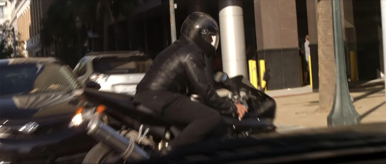 Rage (2014) movie still: medium — A city street with a multi-story building and parked cars in the background. A person on a black sports motorcycle…