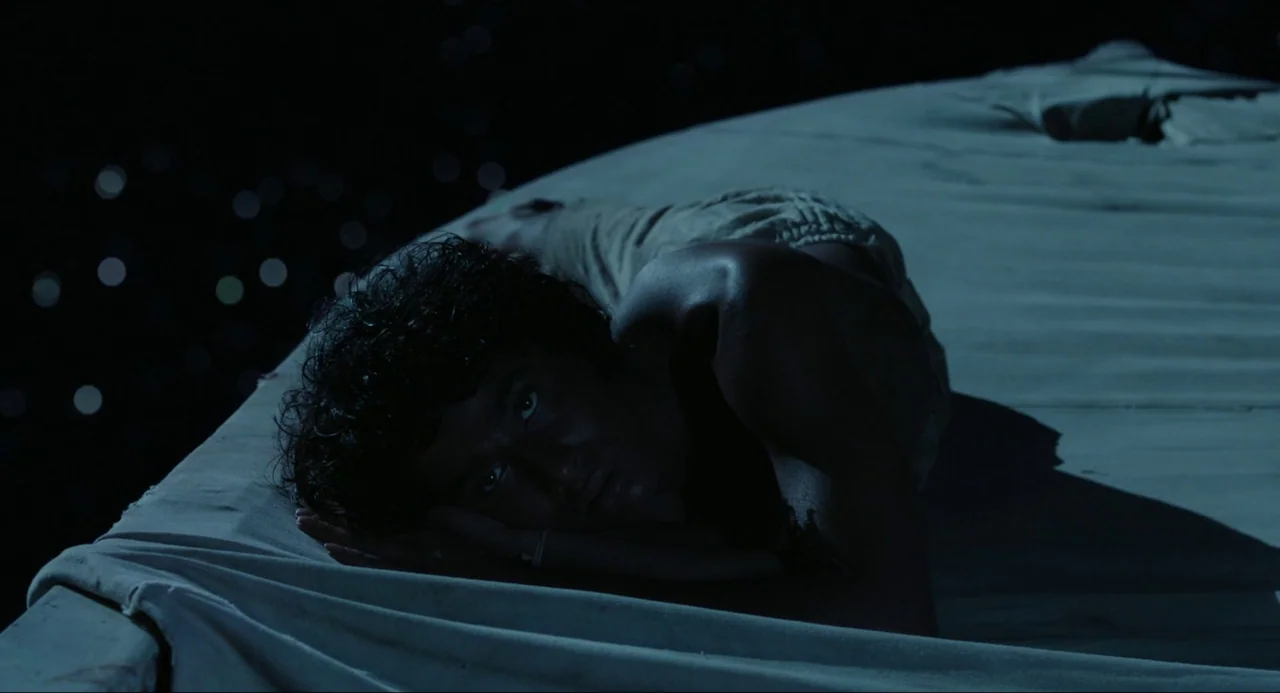 Life of Pi (2012) movie still: medium — A man lies on a wooden raft floating on a dark, calm sea at night. The vast, starry sky creates a backdrop…