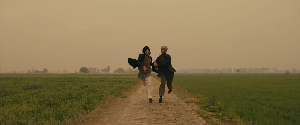 Sardar Udham (2021) movie still: fullbody — A rural dirt path cuts through open, flat green agricultural fields under a hazy sky. Two men in turbans…