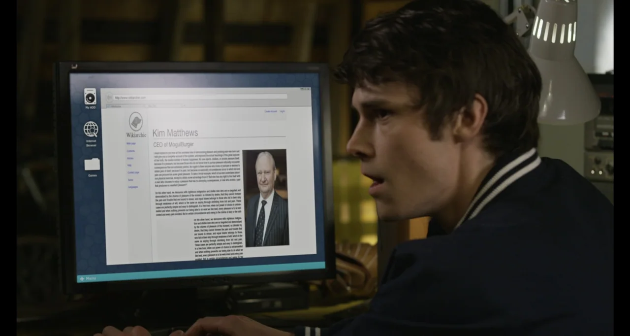 A.R.C.H.I.E. (2016) movie still: medium — A dimly lit room contains a computer desk with a flat-panel monitor displaying data. A young man sits in…