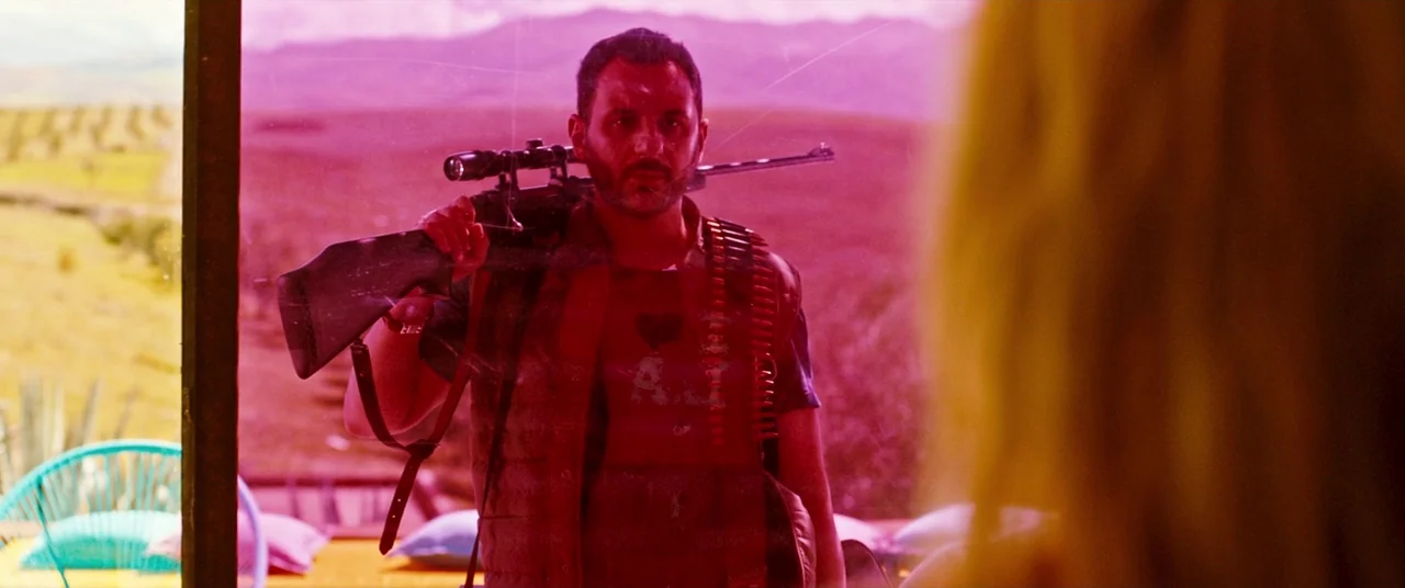 Revenge (2017) movie still: medium — An outdoor patio is visible through a reflective glass pane tinted magenta. A man in a red vest with a rifle…