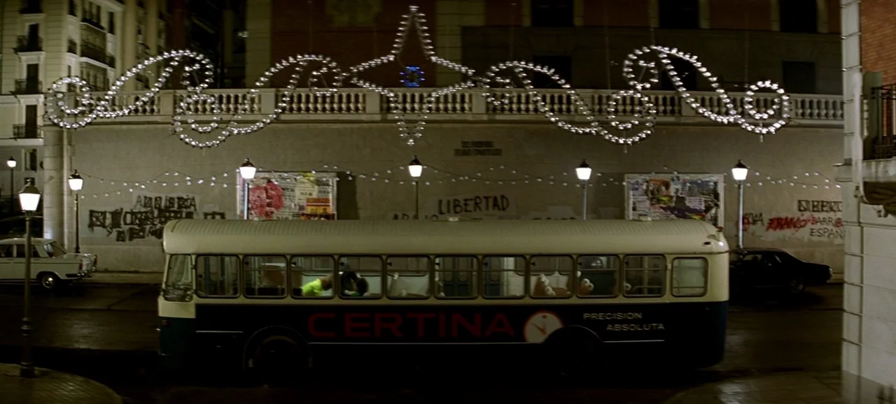 Live Flesh (1997) movie still: wide — An urban street at night features a public bus passing a stone wall. Decorative star-shaped string lights hang…