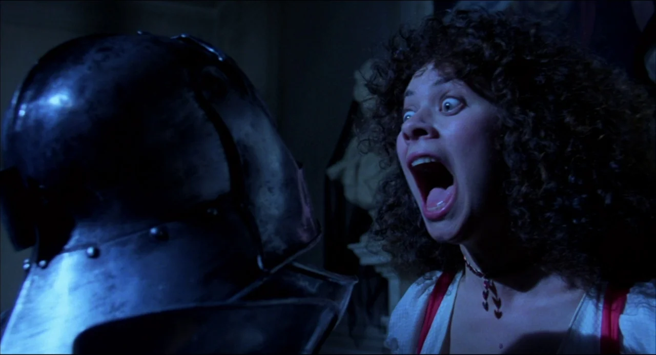 Gothic (1986) movie still: medium — A dimly lit interior space features a dark, reflective metal helmet in the foreground. A woman with curly hair…