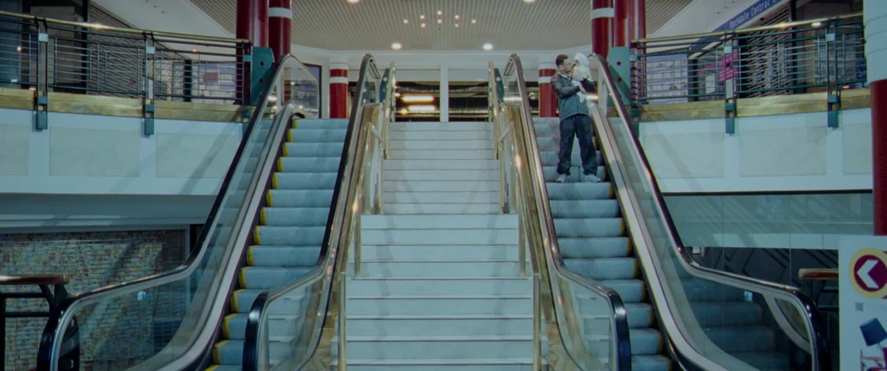 Catch Me Daddy (2014) movie still: wide — A quiet shopping mall interior features symmetrical escalators flanking a central stone staircase. A man…