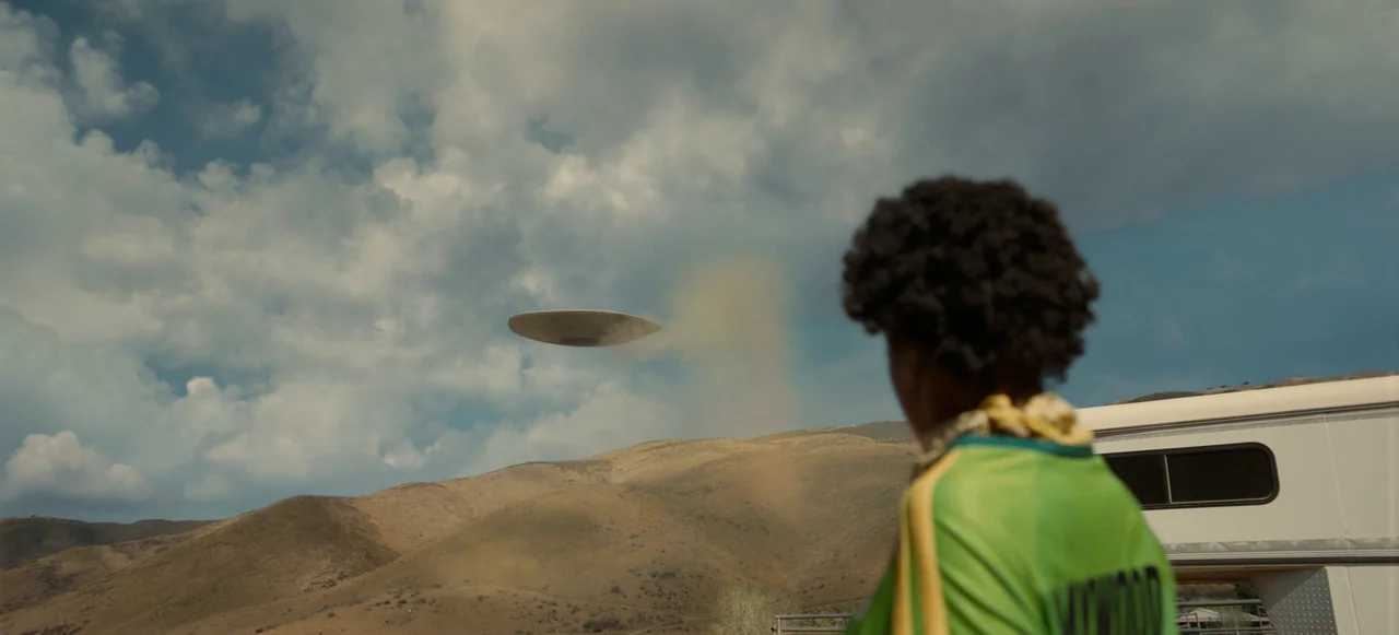Nope (2022) movie still: medium — Dry hills sit beneath a cloudy sky during the day. A person looks toward a metallic disc hovering in the distance…