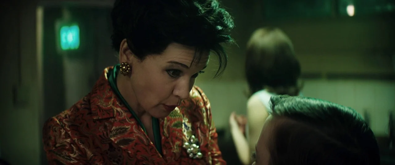 Judy (2019) movie still: closeup — A dimly lit room bathed in green light features an older woman observing someone. She leans forward while wearing…