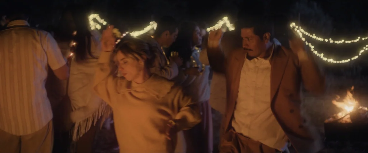 Sign Here (2017) movie still: medium — An outdoor night scene features a group gathered around a fire with string lights. A man and woman dance…