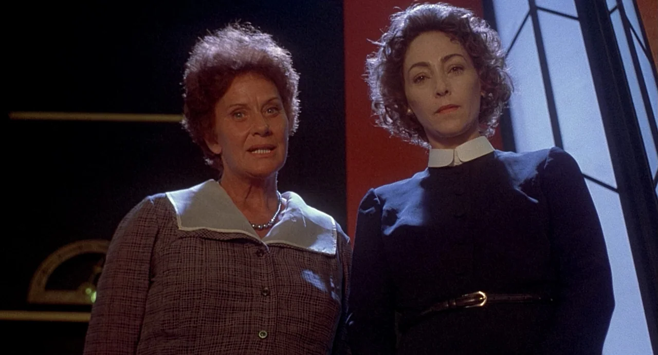 Inferno (1980) movie still: medium — Two women stand in a dark room with a red wall and black background. The older woman on the left stares down…