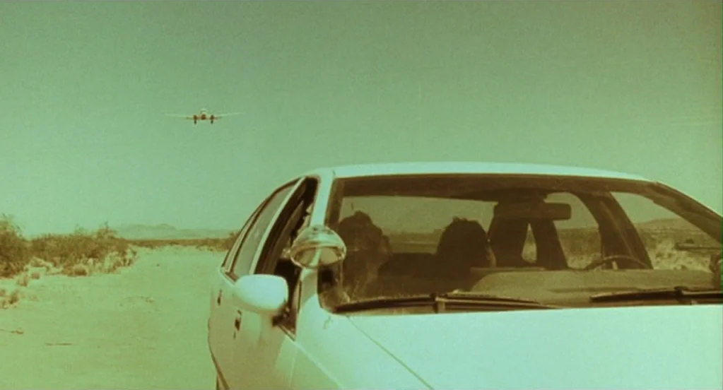 Traffic (2000) movie still: medium — A white sedan is parked on a dusty road in a barren desert landscape. A small airplane is visible in the…