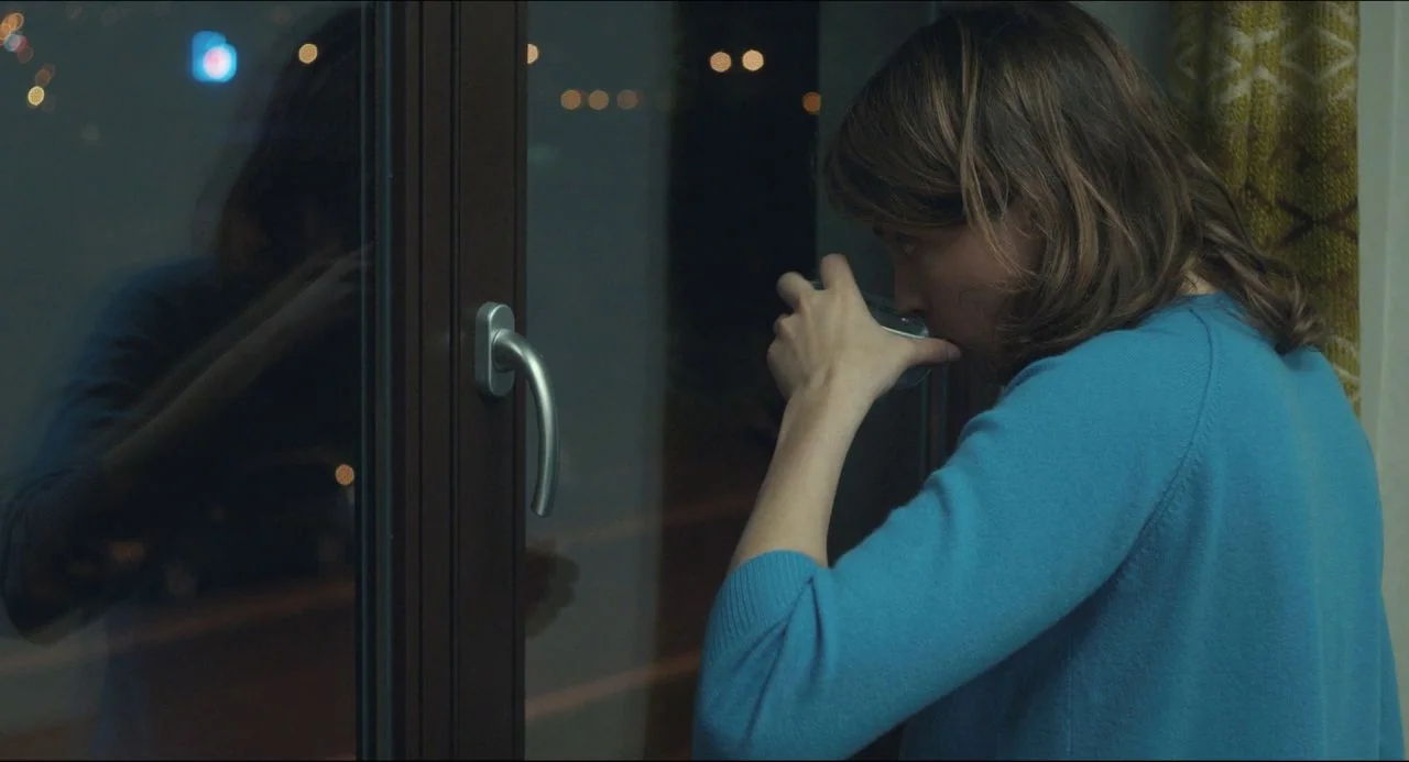 The Unknown Girl (2016) movie still: medium — A dimly lit room with a dark window frame and a patterned curtain visible. A woman stands in profile,…