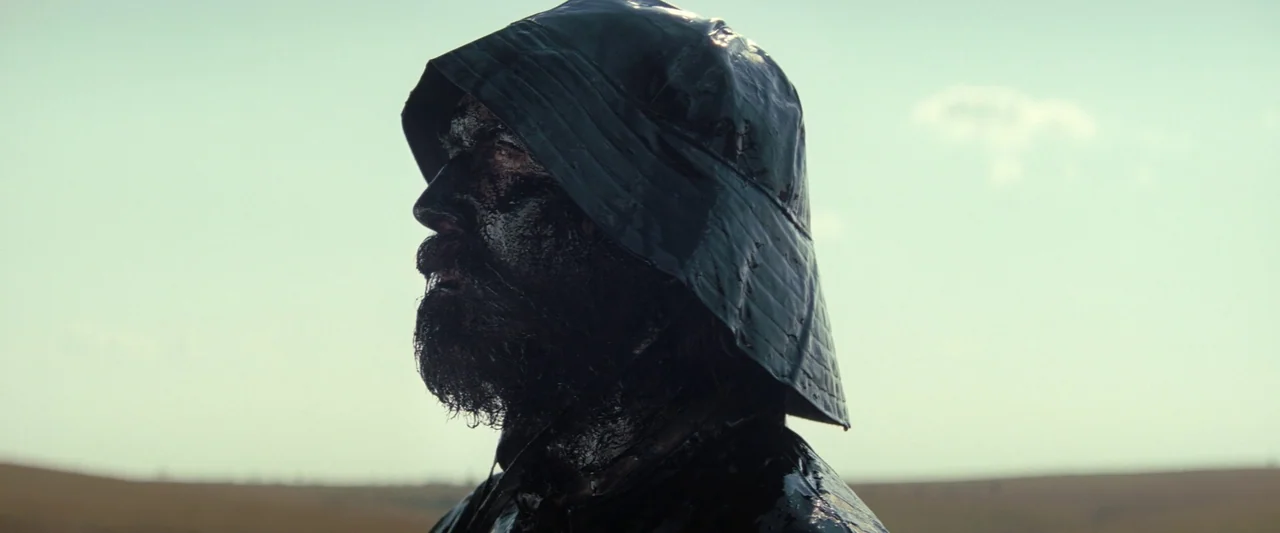 Killers of the Flower Moon (2023) movie still: closeup — A man stands in a desolate, open field under an overcast, bright sky. Thick black oil coats…
