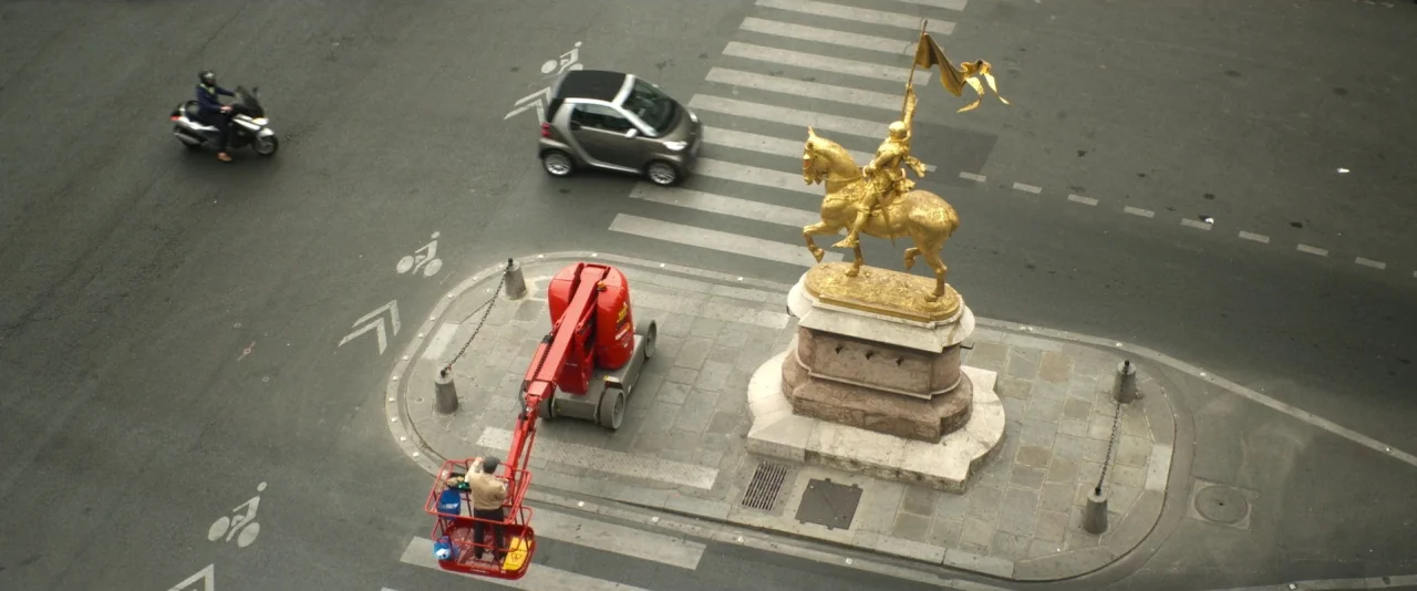 Nocturama (2016) movie still: establishing — A city intersection features a prominent gold equestrian statue on a stone plinth. A worker in a red…
