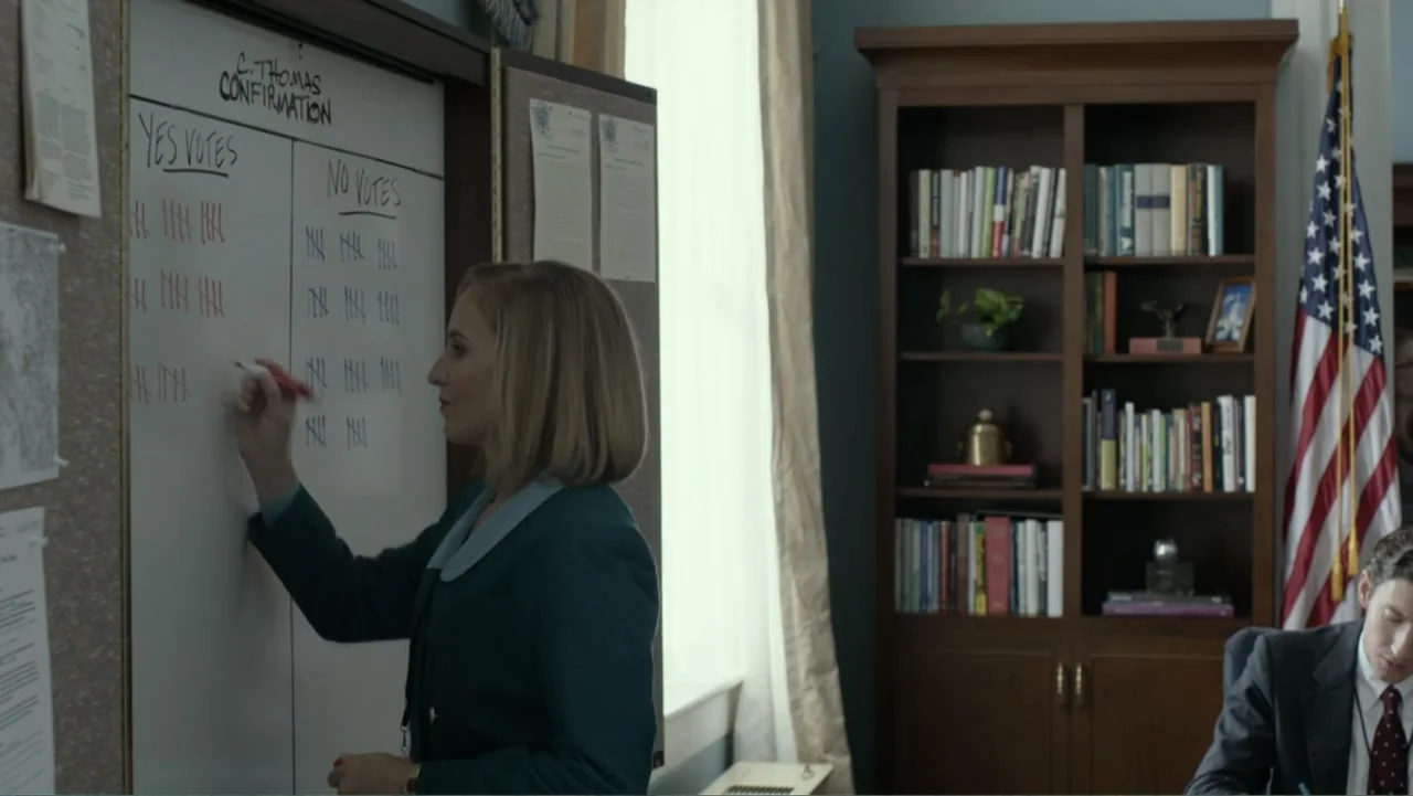 Confirmation (2016) movie still: medium — A bright office contains a wooden bookcase, an American flag, and a whiteboard with tallies. A woman in a…