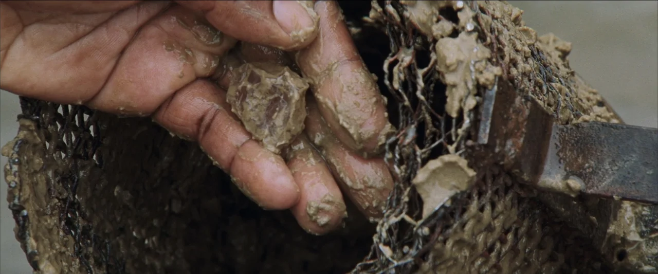 Blood Diamond (2006) movie still: closeup — A close-up view shows human hands holding a small, wet stone coated in mud. A coarse, wire-mesh…