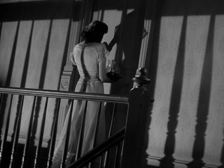 Shadow of a Doubt (1943) movie still: medium — A dimly lit stair landing features patterned wallpaper and a wooden banister. A woman stands by the…