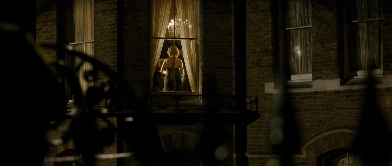 The Oxford Murders (2008) movie still: medium — A brick building facade at night features a glowing window on an upper floor. A woman in a dark…