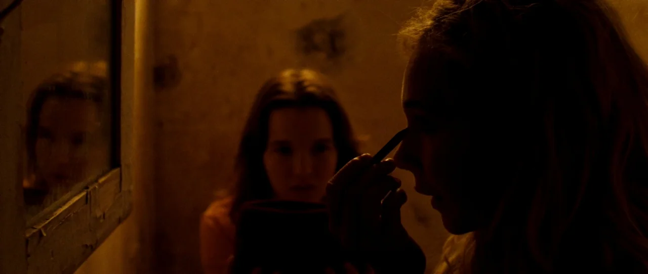 Little Birds (2011) movie still: medium — A dimly lit room with peeling wall texture and a rectangular mirror frame. A woman applies makeup while…
