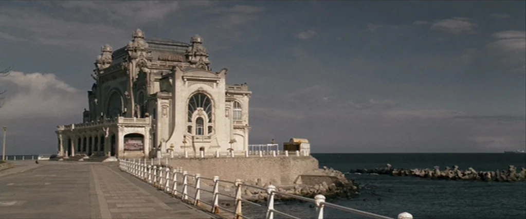 The Brothers Bloom (2008) movie still: establishing — An ornate Art Nouveau building stands on a concrete pier overlooking the sea. Dark clouds fill…