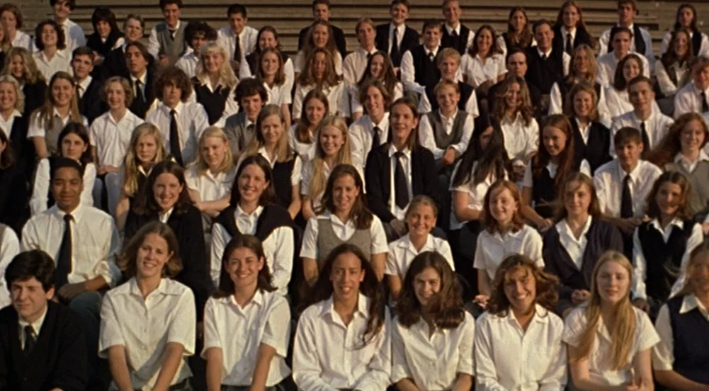 The Virgin Suicides (1999) movie still: wide — A large group of students in uniforms sits on concrete outdoor stadium bleachers. They are organized…