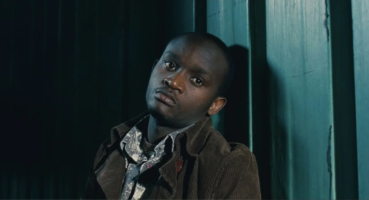 Le Havre (2011) movie still: medium — A narrow indoor corridor features dark, vertical panels on one side. A young man wearing a corduroy jacket…