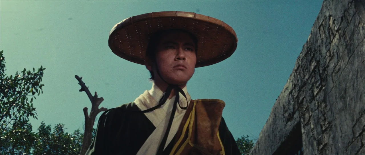 A Touch of Zen (1971) movie still: medium — An outdoor rural setting features a stone wall and sparse foliage against a blue sky. A young man…