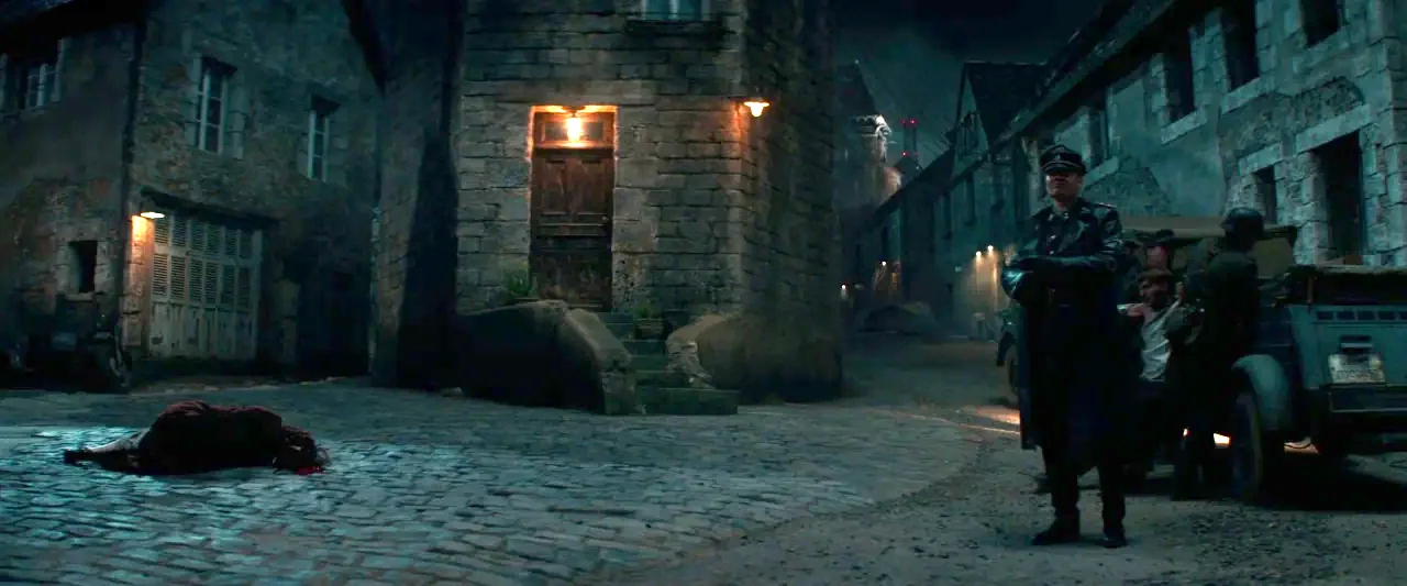 Overlord (2018) movie still: wide — A dimly lit European cobblestone intersection at night features aged stone buildings and low light. A man in a…