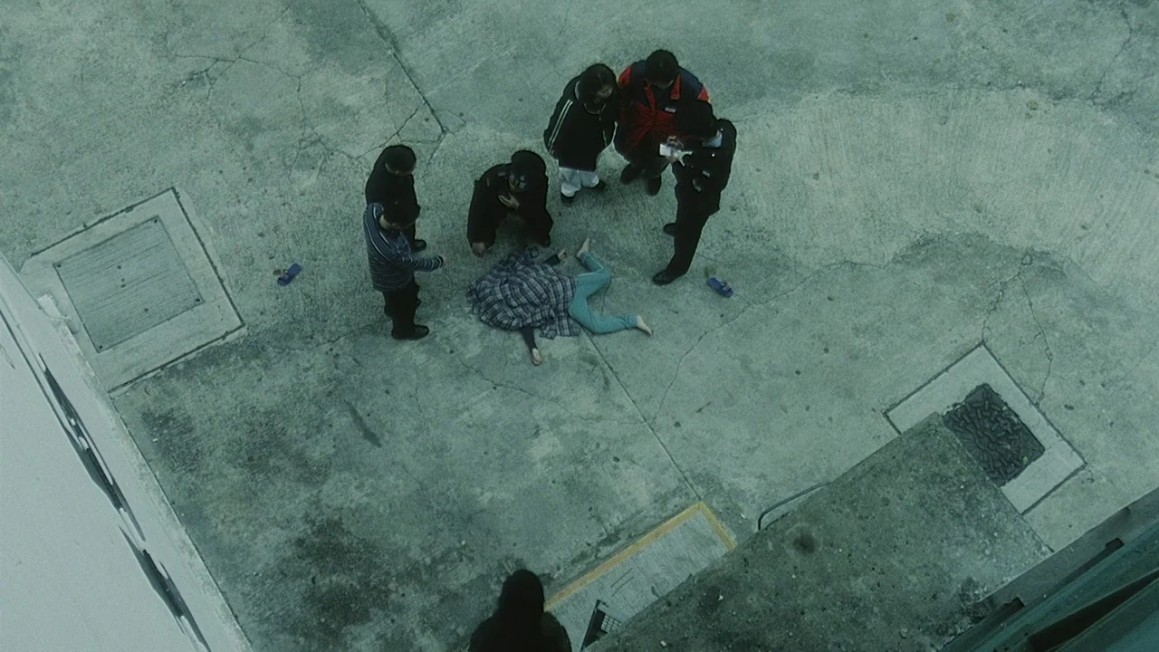 Visible Secret (2001) movie still: wide — A group of people stands in a concrete courtyard around a prone figure. They observe the person lying on…