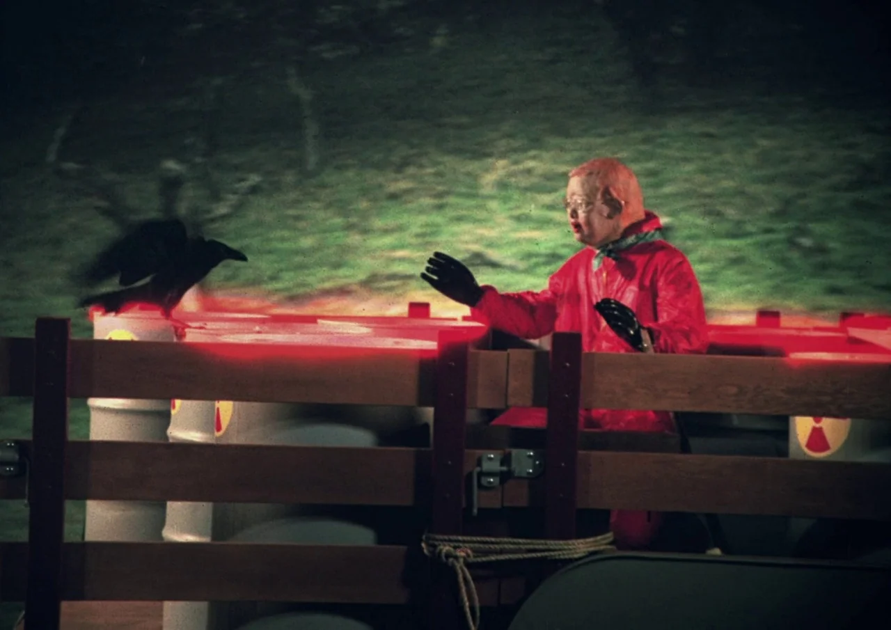 Human Highway (1982) movie still: medium — A dark night setting featuring a wooden fence and barrels marked with radiation symbols. A man in a red…