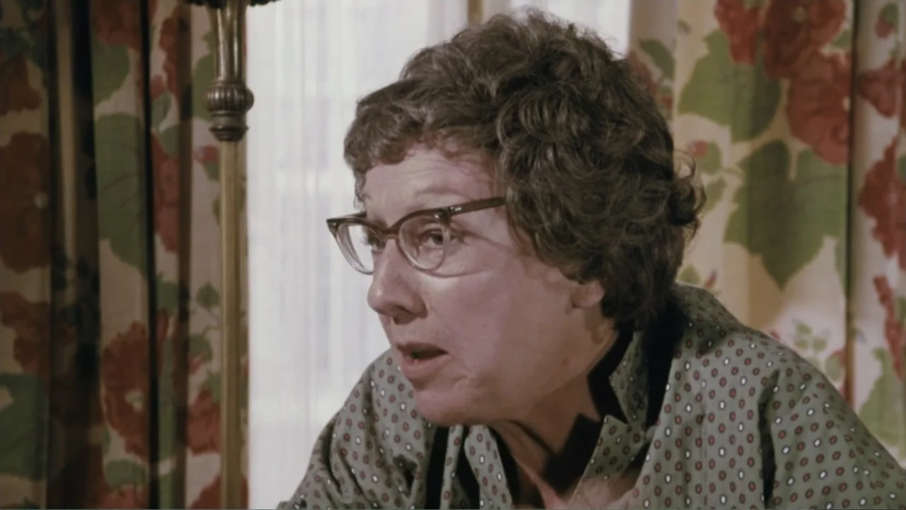 Aunt Mary (1979) movie still: medium — A living room interior features floral curtains with red flowers and green foliage patterns. An older woman…