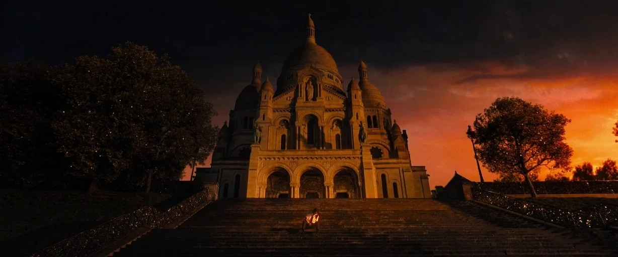 John Wick: Chapter 4 (2023) movie still: wide — A large stone basilica stands at the top of wide stairs under a sunset sky. A solitary figure sits…