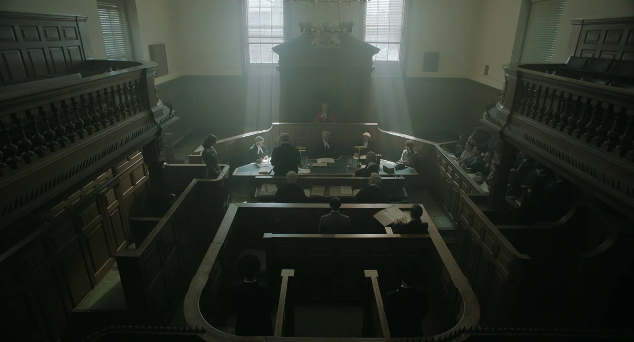 My Policeman (2022) movie still: wide — A wood-paneled, historic room features high-back benches and tiered seating for attendees. Participants in…