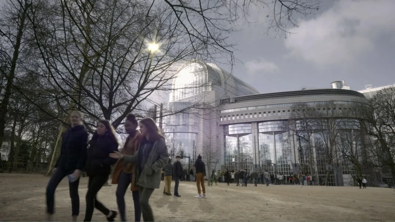 Brexit: Behind Closed Doors (2019) movie still: wide — A group of young people walks across a dirt plaza near a glass modern structure. Bare trees…