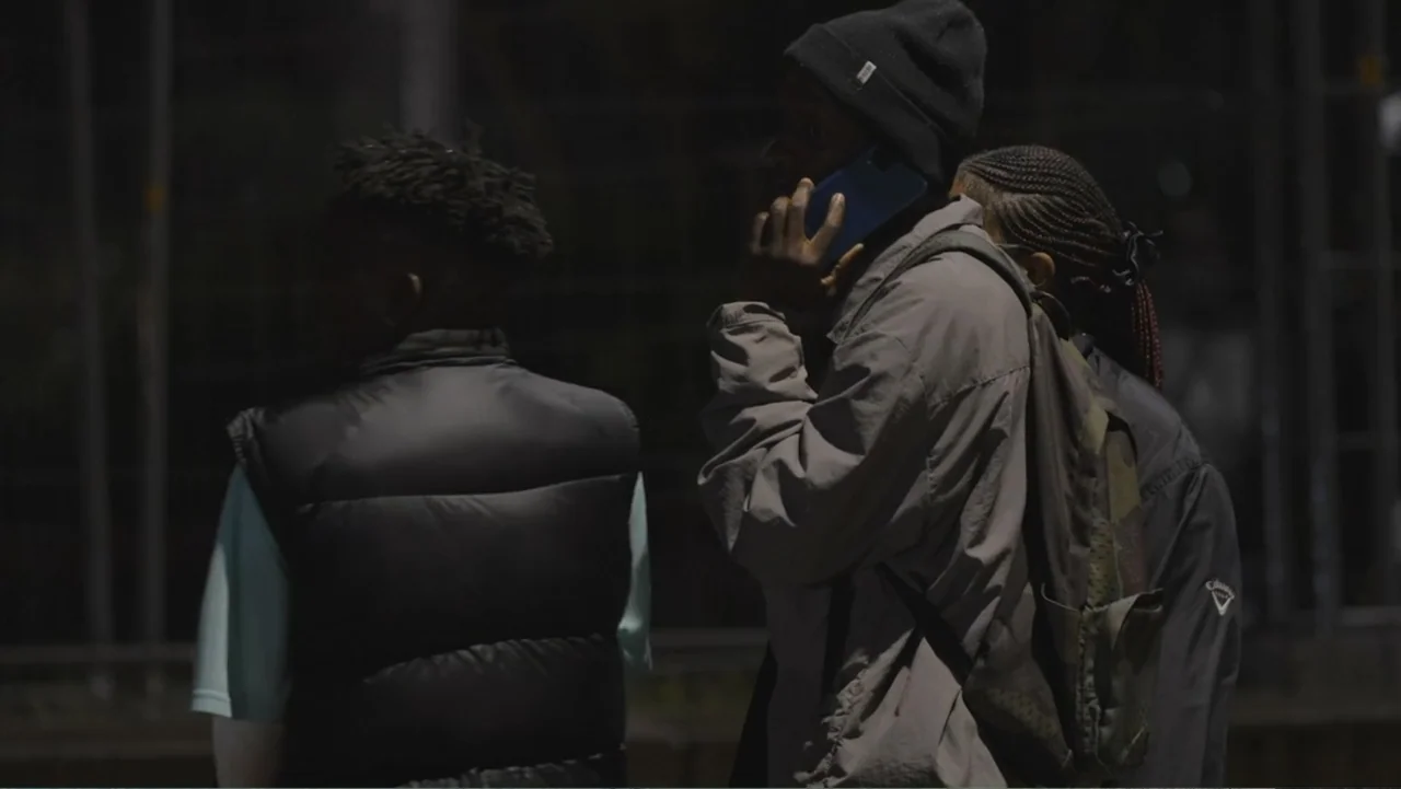 African Throne World Tour (2024) movie still: medium — Three individuals stand together outdoors in bright daylight with a dark fence in the…