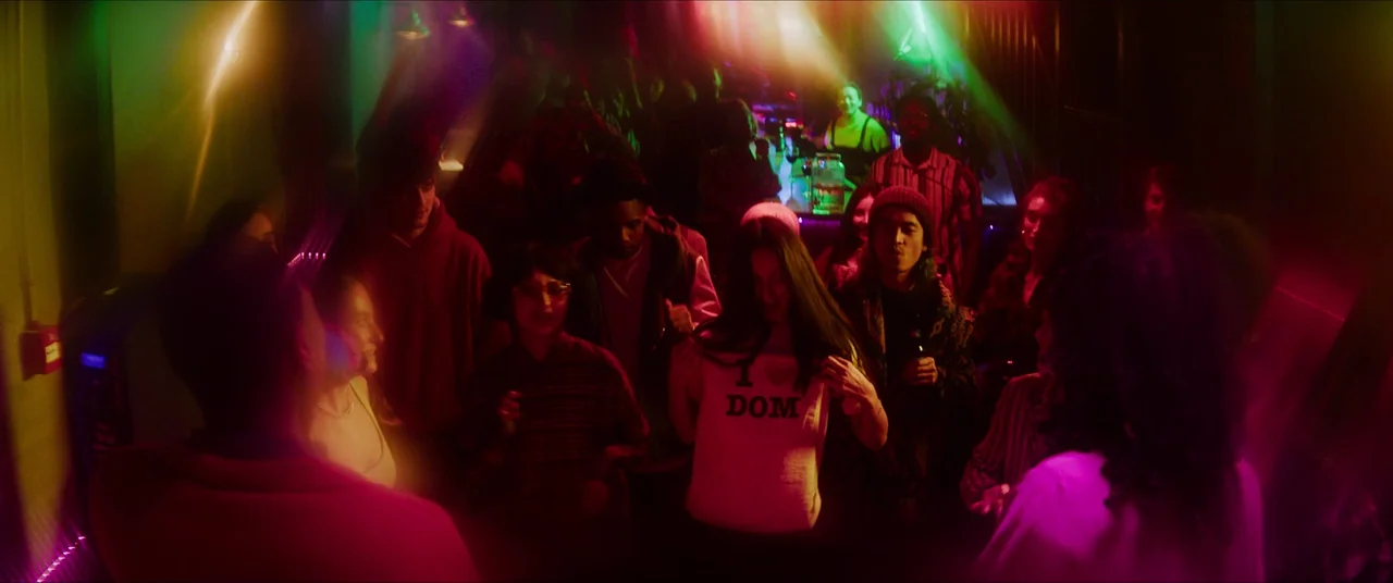 Rye Lane (2023) movie still: medium — A crowded house party interior with low light and colorful neon overhead flares. A group of young adults…