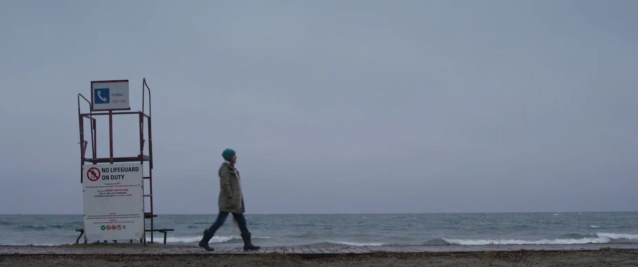 Room (2015) movie still: wide — A wooden boardwalk extends along a grey coastline under a heavy, overcast sky. A woman walks past a tall, metal…