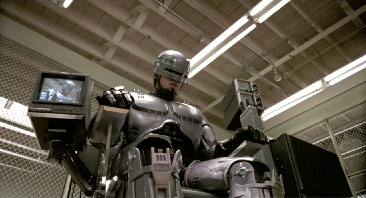 RoboCop (2014) movie still: medium — A high-tech industrial space features white wire mesh partitions and overhead tube lighting. A cyborg sits in a…