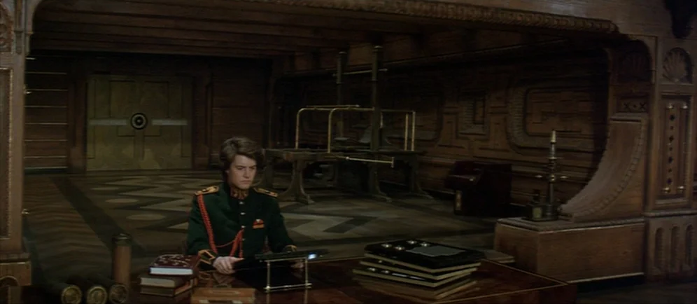 Dune (1984) movie still: wide — A dark, wood-paneled room features intricate carvings and a desk with various books. A man wearing a dark military…