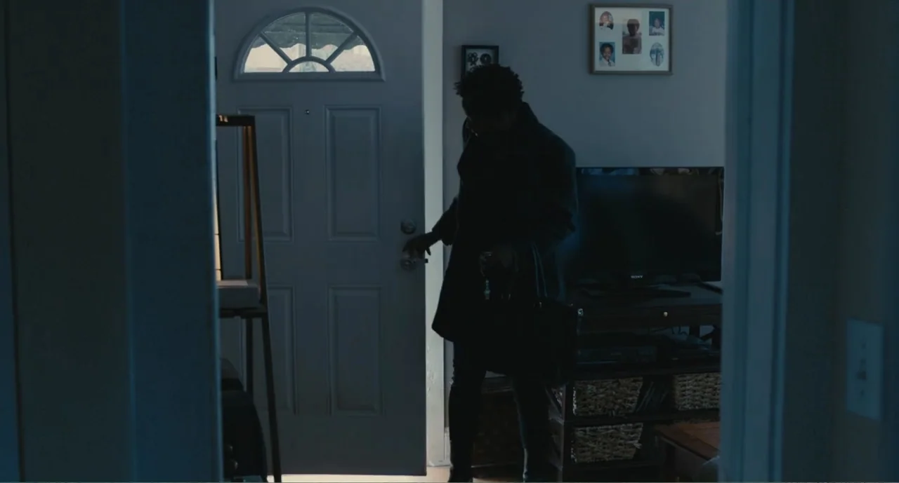 Lila & Eve (2015) movie still: medium — A dark, residential interior space with a television and picture frames on the walls. A man stands in the…