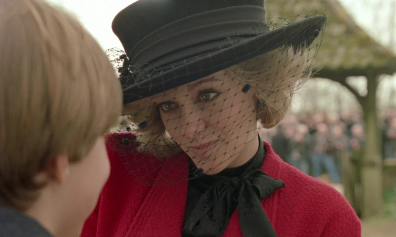 Spencer (2021) movie still: closeup — A woman wearing a mourning hat stands in a churchyard near an obscured crowd. She looks down toward a young…