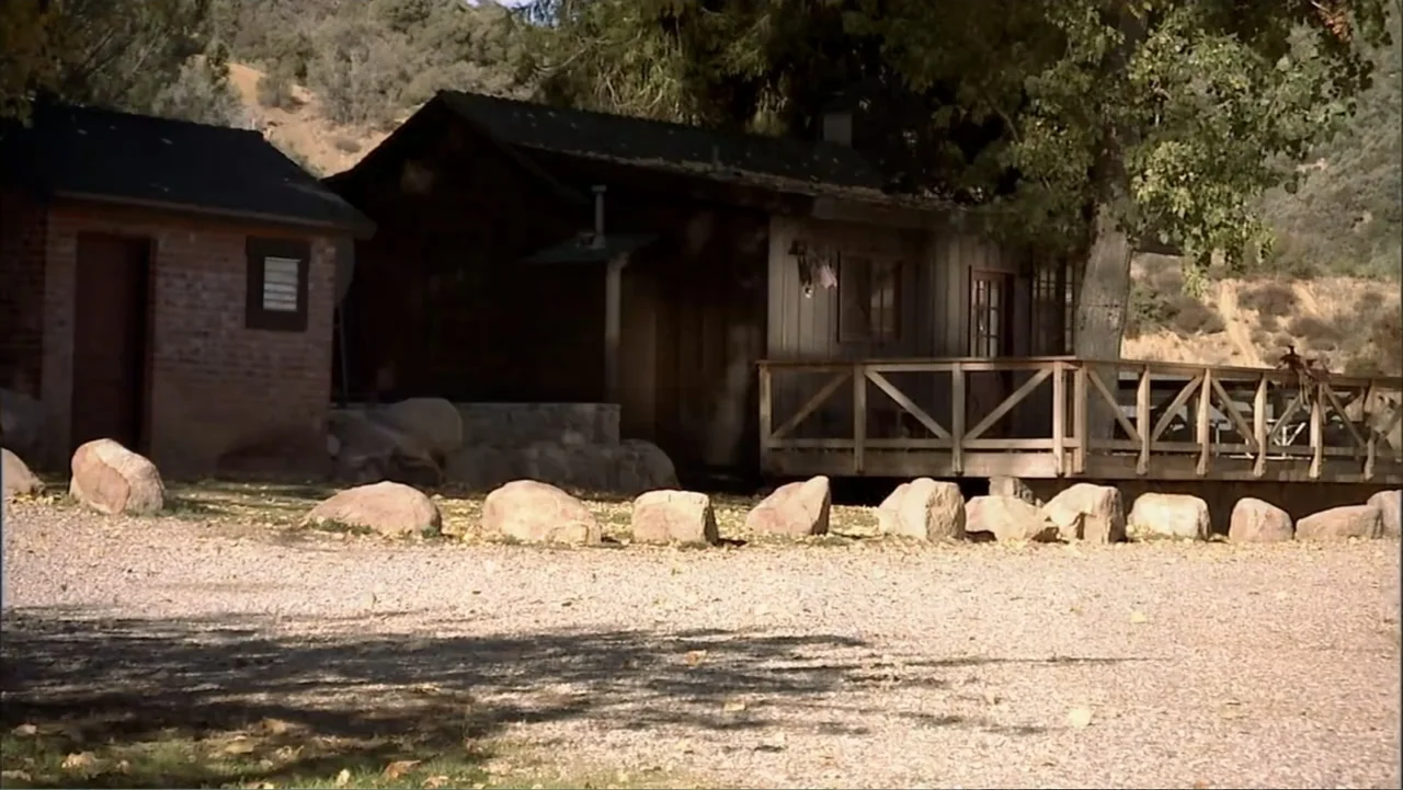 Hot Gimmick: Girl Meets Boy (2019) movie still: wide — A wooden cabin with a porch sits beside a small brick outbuilding. A gravel yard with…