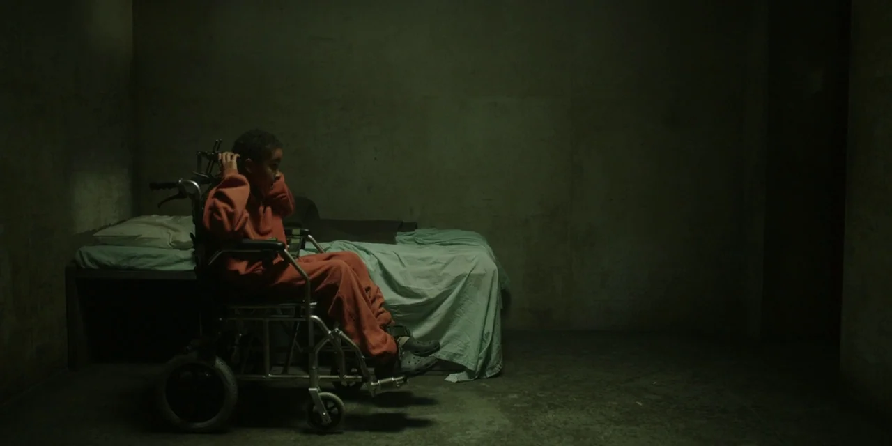 The Girl With All The Gifts (2016) movie still: wide — A stark room with concrete walls features a bed against the back wall. A person in an orange…