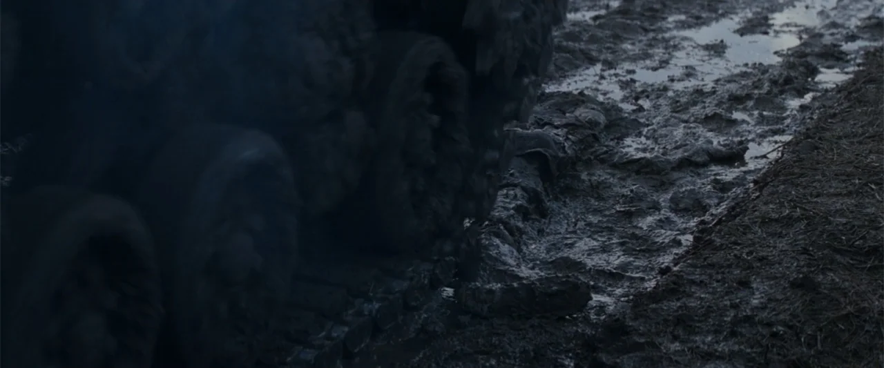 Fury (2014) movie still: closeup — A close-up view of heavy metal tracks rolling over dark, wet, uneven mud. The earth churns and displaces under…