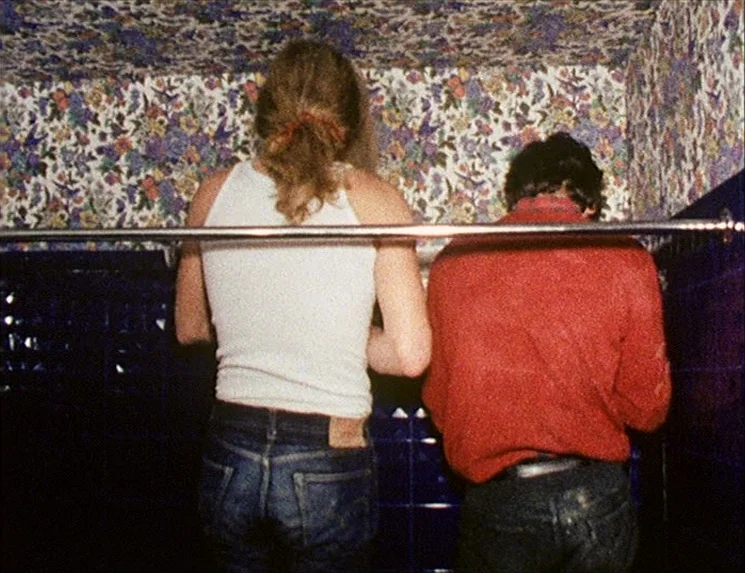 Rio Das Mortes (1971) movie still: medium — A cramped restroom is lined with dark blue wall tiles and floral wallpaper above. Two young men stand…