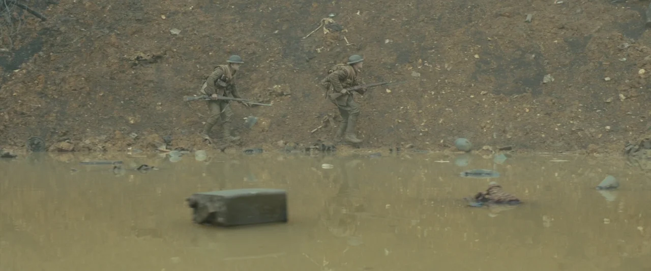 1917 movie still: Two soldiers in period-accurate WWI uniforms trek cautiously along the edge of a stagnant, murky shell-hole. The scene conveys a…