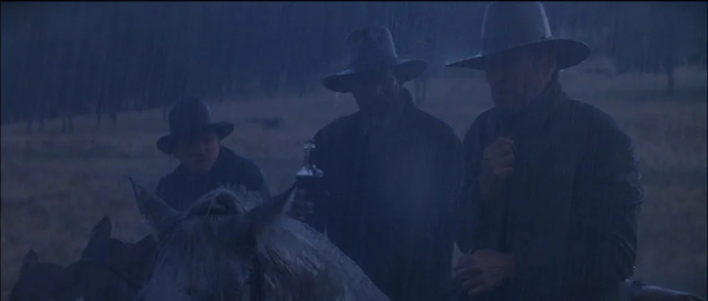 Unforgiven (1992) movie still: medium — Three men in wide-brimmed hats ride horses through a dark, rain-swept field. They move forward in the heavy…