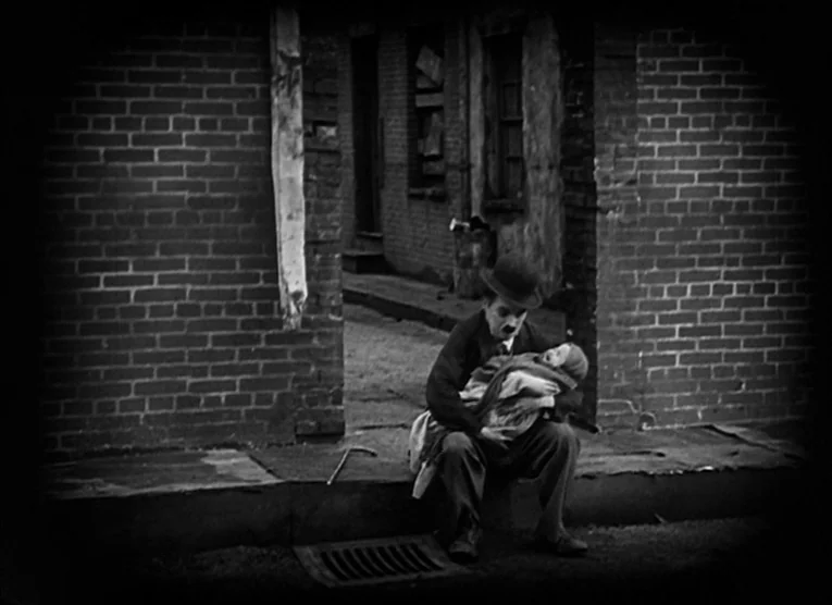 The Kid (1921) movie still: wide — A brick-walled alley entrance features a paved sidewalk and a dark iron drain grate. A man in a suit and bowler…
