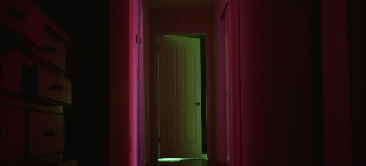 Hellbreeder (2004) movie still: wide — A dark narrow hallway is illuminated by contrasting red and green light sources. An off-center door stands…