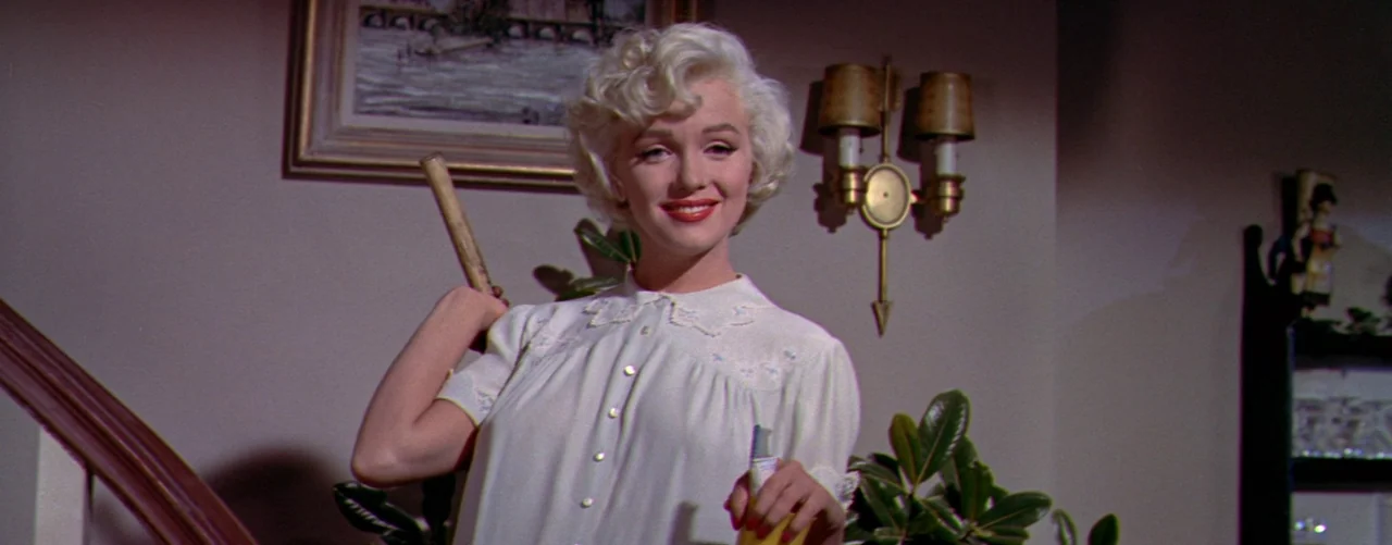 The Seven Year Itch (1955) movie still: medium — A domestic hallway features a wall sconce and a framed print. A blonde woman holds a wooden…
