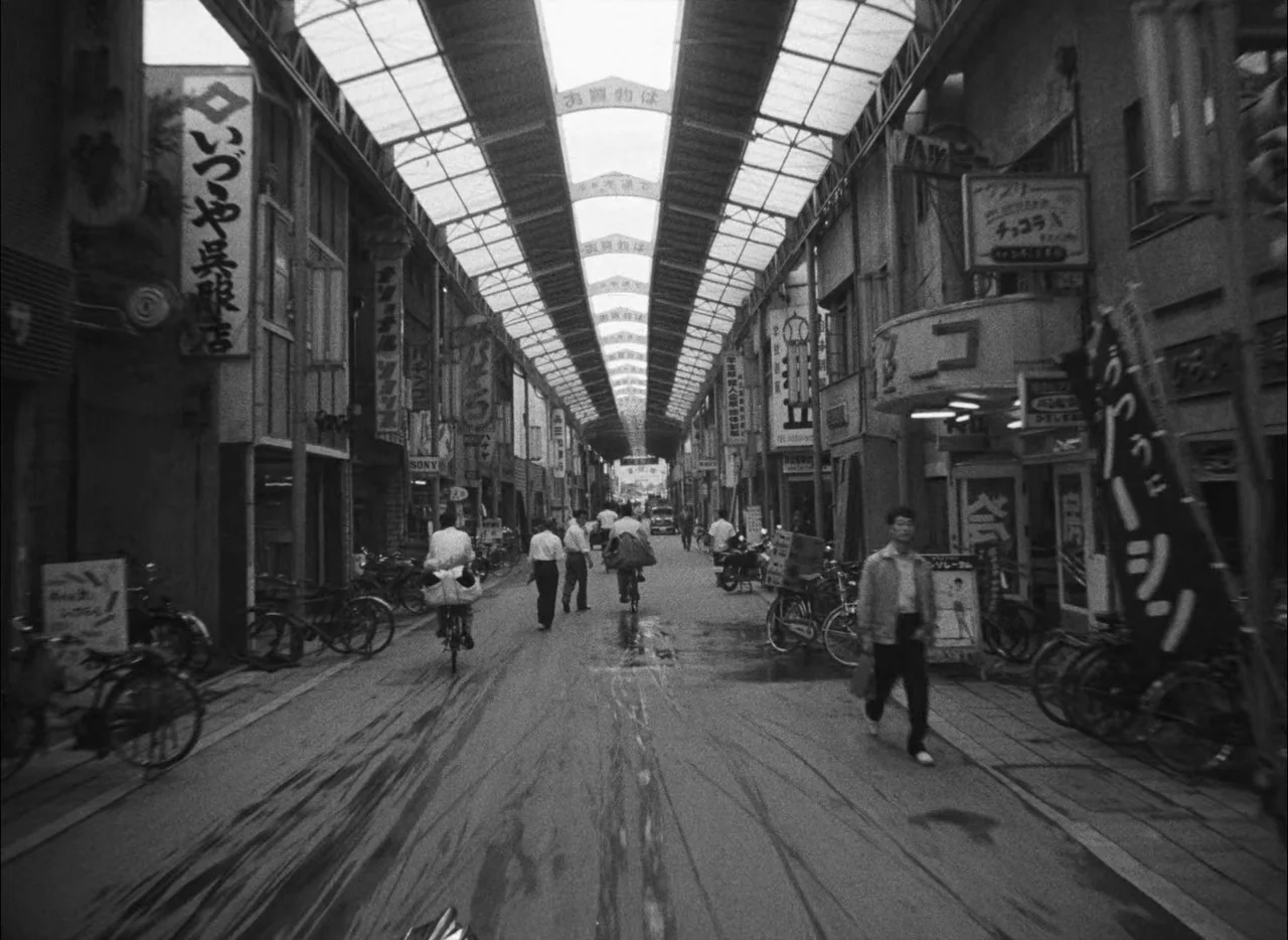 Hiroshima Mon Amour (1959) movie still: wide — A Japanese shopping street is covered by a high, arched translucent roof structure. Pedestrians walk…